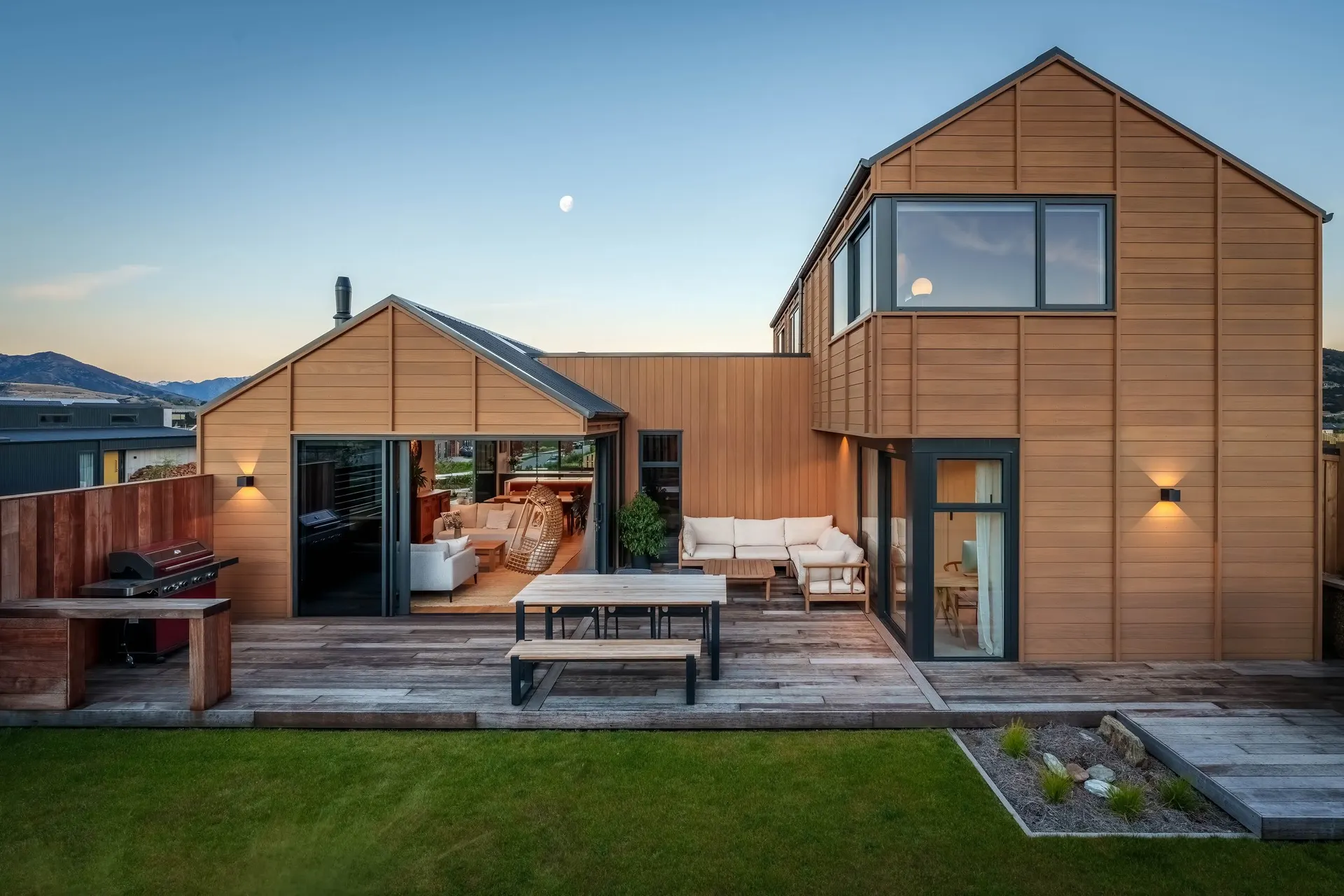 Timber-clad home at dusk, with an outdoor deck, dining table and lounge beside a lawn